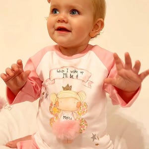 Pu&ograve; includere: Una maglietta a maniche lunghe rosa e bianca con un'immagine di una fata con una corona e ali. La maglietta ha il testo "When I wake up I'll be!" sul davanti. La fata ha il nome "Marcu" scritto sul suo vestito.
