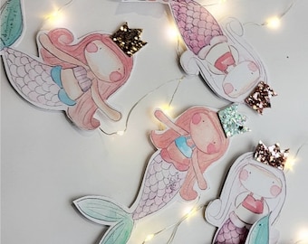 Può includere: Una ghirlanda di luci con sagome di sirene. Ogni sirena ha una coda rosa, blu o verde e una corona. Le sirene sono decorate con glitter e hanno uno stile fantasioso e da cartone animato.