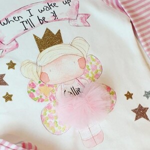 Pu&ograve; includere: Un body bianco con un design rosa e oro che presenta una fata con una corona, ali e un tut&ugrave; rosa. Il body ha il testo "When I wake up I'll be 3!" e il nome "Allie" scritto su di esso.