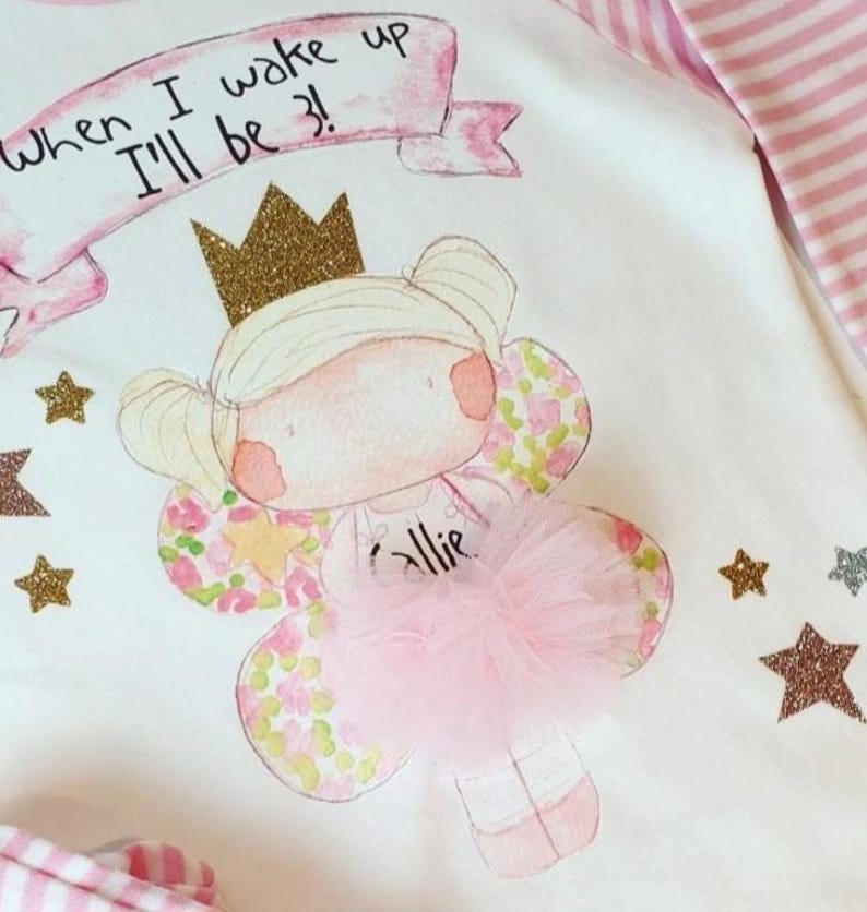 Pu&ograve; includere: Un body bianco con un design rosa e oro che presenta una fata con una corona, ali e un tut&ugrave; rosa. Il body ha il testo "When I wake up I'll be 3!" e il nome "Allie" scritto su di esso.