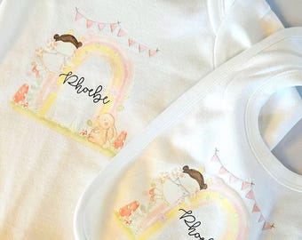Può includere: Set body e bavaglino bianchi per neonati con design ad acquerello. Il design include un arcobaleno, un coniglietto e il nome "Phoebe" in corsivo. Un piccolo banner con bandierine rosa è sopra l'arcobaleno.