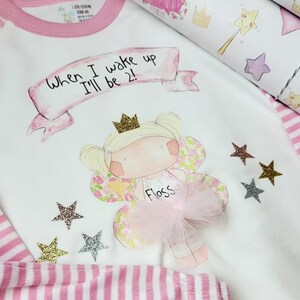 Pu&ograve; includere: Pigiama bianco con strisce rosa e bordino rosa. La parte superiore presenta una fata dei cartoni animati con una corona e il testo "When I wake up I'll be 2!" e il nome "Floss".
