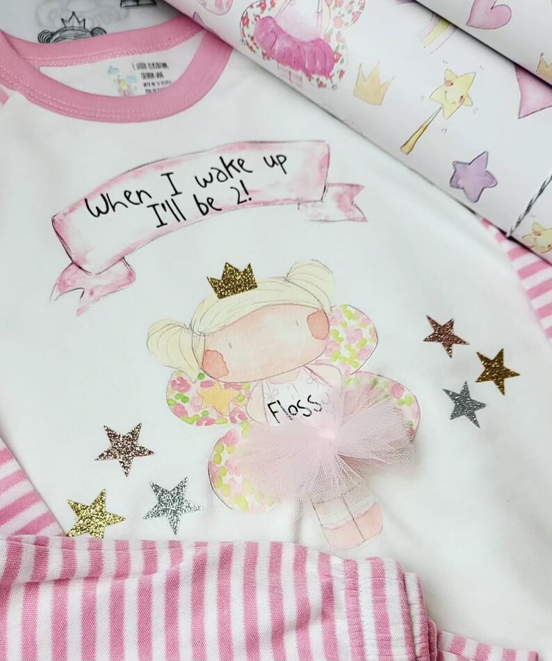 Pu&ograve; includere: Pigiama bianco con strisce rosa e bordino rosa. La parte superiore presenta una fata dei cartoni animati con una corona e il testo "When I wake up I'll be 2!" e il nome "Floss".