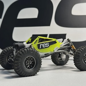 May include: A small, off-road RC car with a bright yellow roll cage and large, black tires. The car has the word "res" printed on the side. The suspension features orange springs and the chassis is white and silver.