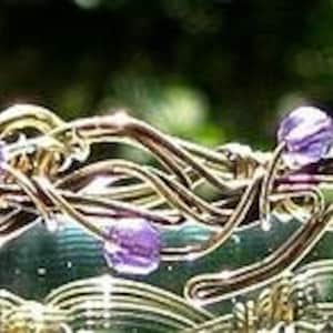 Może przedstawiać: Bransoletka ze złotego drutu z fioletowymi szklanymi koralikami. Drut jest skręcony w wzór przypominający winorośl.