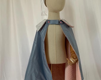 Könnte beinhalten: Ein umkehrbarer Umhang in Blau und Rosa mit einer spitzen Kapuze. Der Umhang hat einen weißen Saum und zwei rosa Sternknöpfe. Die Kapuze ist mit weißer Spitze gefüttert.