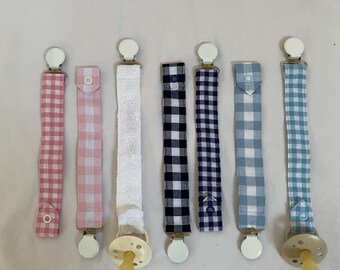 Könnte beinhalten: Eine Sammlung von Schnullerclips für Babys in verschiedenen Mustern. Die Clips haben Stoffbänder in rosa Vichy, weißer Lochstickerei und blauem Vichy-Design. Jeder Clip hat einen weißen Kunststoffclip und einen passenden Schnuller.