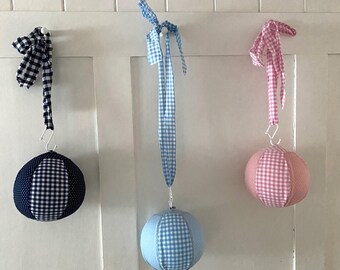 Könnte beinhalten: Drei Stoffbälle mit Gingham-Mustern in Blau, Rosa und Weiß, die an Haken an einer weißen Wand hängen. Die Bälle sind an Bändern mit Schleifen befestigt.