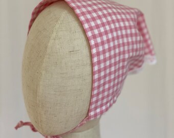 Könnte beinhalten: Eine rosa-weiße Gingham-Haube mit weißer Spitzenborte. Die Haube ist mit einem rosa Band gebunden.