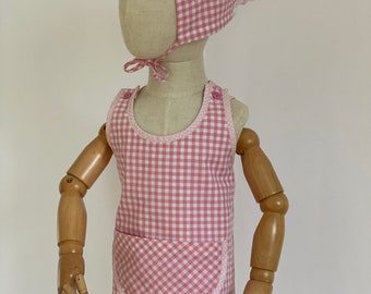 Könnte beinhalten: Rosa-weißes Gingham-Schürzenkleid mit großer Tasche und passender Haube. Die Haube hat einen weißen Spitzenbesatz.