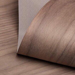 May include: A close-up of a sheet of wood veneer with a light brown color and a wood grain pattern. The veneer is folded over on itself, revealing a light gray backing.