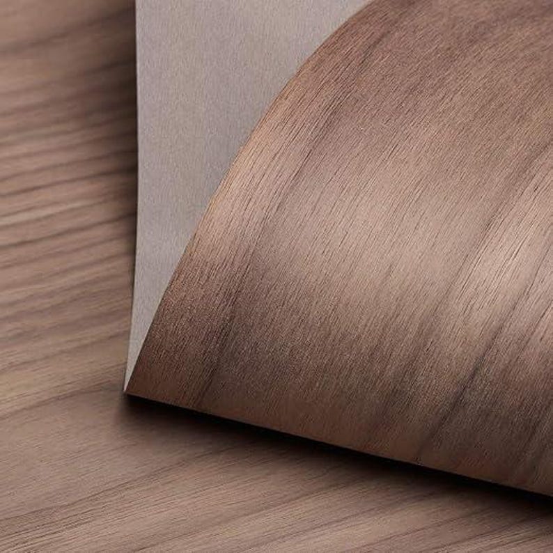 May include: A close-up of a sheet of wood veneer with a light brown color and a wood grain pattern. The veneer is folded over on itself, revealing a light gray backing.