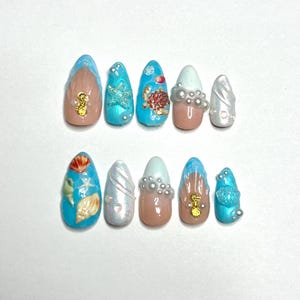 Könnte beinhalten: Ein Set von zehn künstlichen Nägeln mit Meeresmotiven. Die Nägel zeigen Designs von Muscheln, Seesternen und Schildkröten, mit Akzenten in Blau, Gold und Perlmutt. Einige Nägel haben eine nackte Basis, andere sind blau.