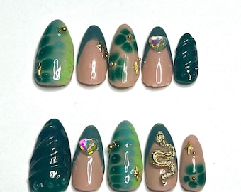 May include: A set of ten artificial nails in various designs. The nails feature a mix of colors, including green, teal, and nude. Some nails have gold accents, rhinestones, and a gold snake design. The nails are oval-shaped.