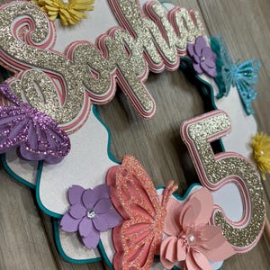 May include: A cake topper with the name "Sophia" in gold glitter letters, a number "5" in gold glitter, and pink, purple, and blue butterflies and flowers.