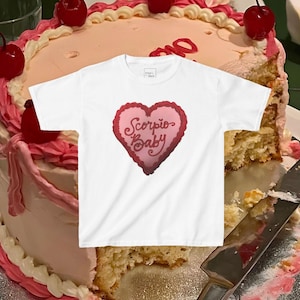 Peut inclure: T-shirt blanc avec un graphique en forme de cœur rose sur lequel est inscrit "Scorpio Baby". Le t-shirt est posé sur un gâteau glacé rose et blanc.