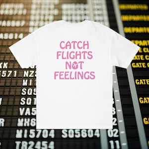 Peut inclure: T-shirt blanc avec l'inscription rose "CATCH FLIGHTS NOT FEELINGS" et un avion. En arrière-plan, un tableau des départs est visible.