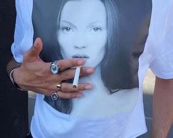 May include: White t-shirt featuring a black and white portrait of a woman. The person is wearing rings and a bracelet. A cigarette is held between the fingers. The person is also wearing a leopard print belt with a gold buckle.