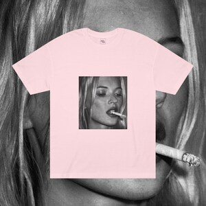Puede incluir: Una camiseta rosa con una imagen en blanco y negro de una mujer fumando un cigarrillo. La mujer tiene el pelo largo y rubio y lleva una camisa blanca.