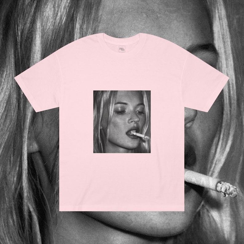 Puede incluir: Una camiseta rosa con una imagen en blanco y negro de una mujer fumando un cigarrillo. La mujer tiene el pelo largo y rubio y lleva una camisa blanca.