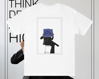 Può includere: Maglietta bianca con una grafica di una persona che tiene un cartello con la scritta "Think Bigger."