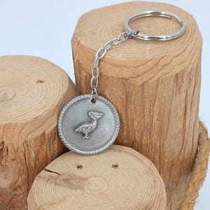 May include: A silver-colored keychain with a pelican emblem hangs from a wooden log display. The display is made of three light brown wooden logs bound together with a rope. The keychain has a circular pendant with a detailed pelican design.