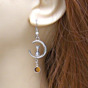 May include: Silver dangle earrings featuring a crescent moon with a cat silhouette and a dangling amber-coloured gemstone.