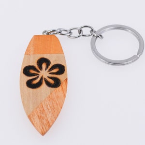 May include: Wooden surfboard-shaped keychain with a black floral design. The keychain is made of light and dark brown wood, with a silver metal keyring and chain. The design is a stylized flower.