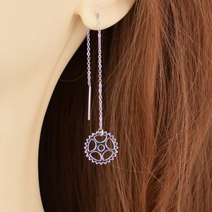 May include: Silver chain earrings with a small gear charm hanging from one chain. The earrings are shown on a model with long brown hair.