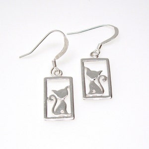 May include: A pair of silver cat earrings. Each earring has a rectangular frame with a cutout silhouette of a cat. The cat is sitting with its tail curled up. The earrings have a hook closure.