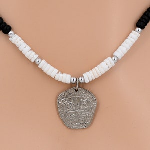 以下が含まれることがあります： 銀貨のペンダントネックレスで、白と黒の貝殻ビーズのチェーンが付いています。コインは複雑なデザインで、人気の海賊の宝のモチーフです。