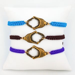 May include: Three bracelets featuring woven cord bands in bright blue, dark brown, and purple. Each bracelet is centered with a metallic bronze-toned shark jaw charm, showcasing detailed teeth and a realistic anatomical shape connected by small gold rings.