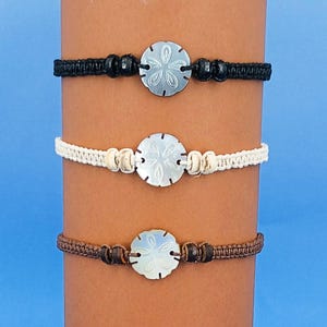 May include: Three macrame bracelets featuring a central sand dollar charm. The top bracelet is black, the middle is off-white, and the bottom is brown. Each bracelet has a woven cord band with two small beads flanking the sand dollar centerpiece.