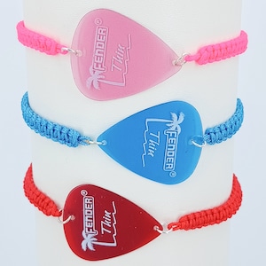 Könnte beinhalten: Drei Armbänder mit Fender Thin Gitarrenplektren als Mittelstücke. Das obere Armband ist neonpink, das mittlere leuchtend blau und das untere rot. Jedes Plektrum ist mit silbernen Metallringen an einem farblich passenden geflochtenen Band befestigt.