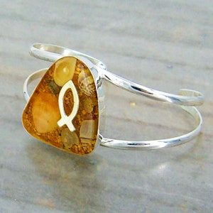 May include: A silver cuff bracelet with a triangular shaped charm. The charm is filled with sand and seashells and features a silver fish symbol.