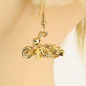 May include: Gold-coloured motorcycle earrings. The earrings feature a detailed miniature motorcycle design, including wheels, handlebars, and a seat. The earrings hang from a gold-coloured hook.