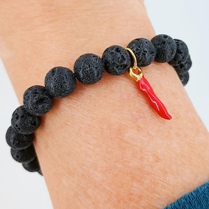 May include: A bracelet featuring black lava rock beads and a red chilli pepper charm. The bracelet has round, textured black beads. The charm is a small, gold-coloured ring holding a red chilli pepper pendant.