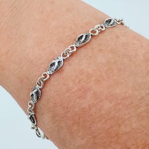 May include: Silver-toned bracelet featuring a series of flip-flop charms. The bracelet is made of small, interconnected links, with each charm displaying detailed flip-flop designs. The bracelet is shown on a person's wrist.