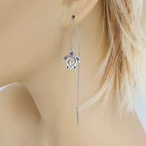 May include: A silver-toned, dangling earring featuring a small turtle charm. The turtle has an openwork design. The earring hangs from a delicate chain that threads through the ear. The earring is shown on a person's ear.