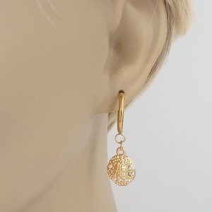May include: Gold-colored hoop earring with a dangling charm. The charm is a detailed, circular design resembling a sand dollar. The earring is attached to the ear. The earring is gold-colored.