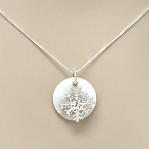 May include: A silver necklace featuring a round, white pendant with a snowflake design. The snowflake is made of silver and is attached to the pendant. The necklace has a delicate chain.