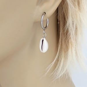 May include: A single silver hoop earring with a white cowrie shell pendant. The shell has a dark vertical stripe. The earring is shown worn on an ear.