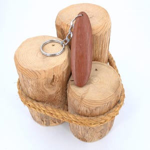 May include: A brown wooden keychain with a silver chain and key ring. The keychain is oval-shaped and rests on three wooden log slices tied together with rope. The logs are light brown with visible wood grain.