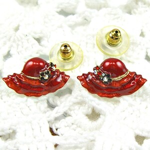 May include: A pair of red hat stud earrings. Each earring features a glossy red hat design with a gold band and a small floral accent with clear and black rhinestones. The earrings have gold-colored posts and clear plastic backs.