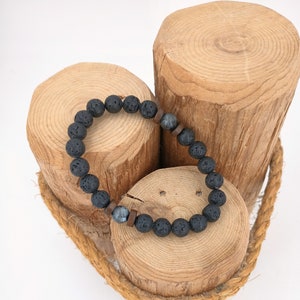 Op de afbeelding: Een armband van zwarte lavasteen kralen met twee grijze en bruine houten kralen. De armband wordt getoond op drie houten blokken.