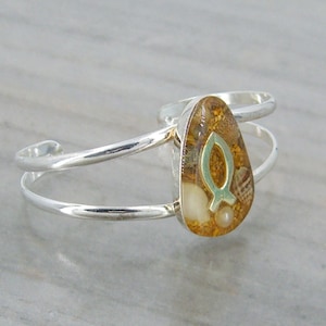 May include: Silver cuff bracelet with a teardrop-shaped charm. The charm is filled with sand and seashells and features a gold-colored fish symbol.