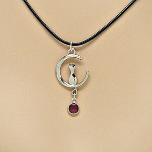 May include: A silver necklace with a crescent moon pendant featuring a cat silhouette. A small, dark purple gemstone dangles from the bottom of the pendant.
