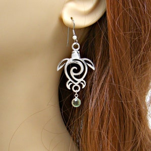 May include: Silver dangle earrings featuring a turtle design with a swirl pattern and a green gemstone drop.