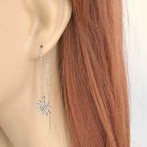 May include: A pair of silver chain earrings. Each earring features a sun-shaped charm with a spiral center and a dangling bar. The earrings are shown on a mannequin with auburn hair.