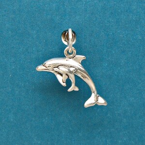 May include: A silver dolphin charm featuring a larger dolphin with a smaller one swimming beside it. The charm has a loop for attaching to jewellery. The dolphin's tail is curved. The charm is set against a blue background.